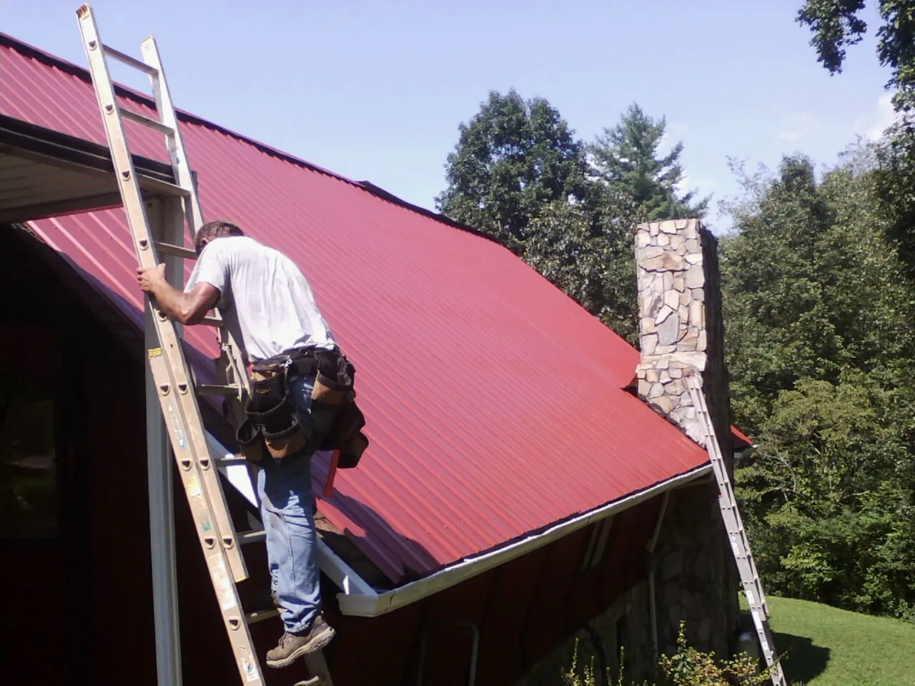 Roofer on a ladder beside a red metal roof and stone chimney — Soffit & Fascia Repair by Roofing Brothers in Cheyney